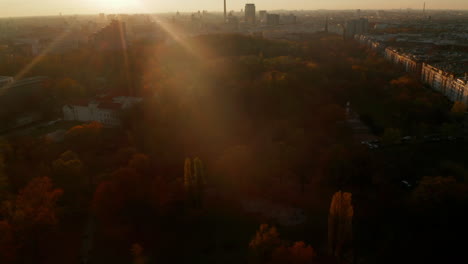 Orange-and-Red-colored-Tree-forest-or-Public-Park-in-Big-City,-Contrast-between-Urban-environment-and-Nature,-Aerial-dolly-out-tilt-up-above-Berlin,-Germany-Cityscape