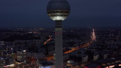 Slowly-lowering-Aerial-Wide-View-of-TV-Tower-Skyscraper-with-Cityscape-at-Night-in-Berlin,-Germany,-Drone-perspective-from-above