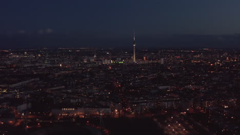 Flight-over-large-city-at-night.-Aerial-view-of-urban-neighbourhood-and-town-dominant,-tall-thin-TV-tower-Fernsehturm.-Berlin,-Germany.