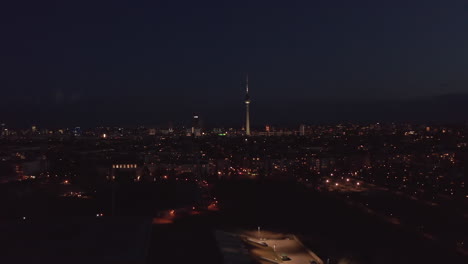 Flight-over-urban-neighbourhood-at-night.-Aerial-view-of-dimly-lit-streets-and-buildings.-Tall-and-thin-TV-tower-Fernsehturm-with-red-blinking-lights.-Berlin,-Germany.