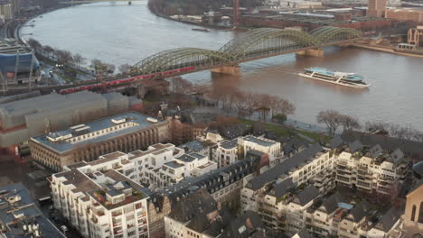 Fly-above-urban-borough.-Tilt-up-reveal-commuter-train-passing-over-wide-Rhine-river-on-bridge.-Cologne,-Germany