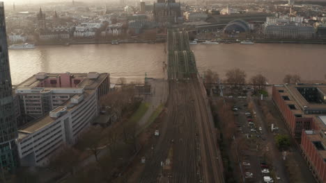 Aerial-view-of-railway-Hohenzollernbrucke-bridge-over-wide-Rhine-river-flowing-through-city.-Tilt-up-reveal-gothic-Cologne-Cathedral-against-sunset-sky.-Cologne,-Germany