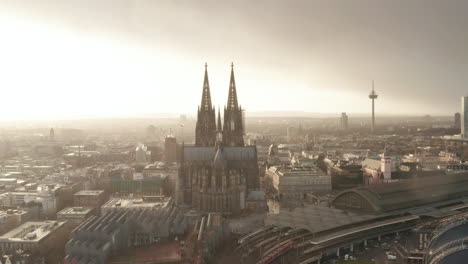 Aerial-shot-of-gothic-Cologne-Cathedral-against-bright-light-sky-in-light-rain.-Tilt-down-on-trains-arriving-or-leaving-train-station.-Cologne,-Germany