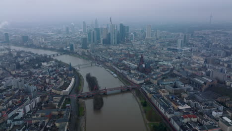 High-angle-view-of-building-in-urban-borough-along-river.-Tilt-up-reveal-hazy-cityscape-with-downtown-skyscrapers.-Frankfurt-am-Main,-Germany