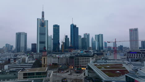 Forwards-fly-above-buildings-around-Rossmarkt.-Group-of-modern-downtown-skyscrapers-against-overcast-sky-at-twilight.-Frankfurt-am-Main,-Germany