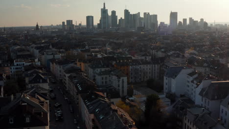 Forward-moving-and-rising-drone-Aerial-view-of-town-neighbourhood-and-skyscrapers-in-distance.-Downtown-skyline-against-sun.-Frankfurt-am-Main,-Germany