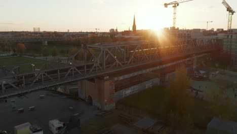 Establishing-Shot-of-Iconic-Berlin-Yellow-Subway-Train-passing-Bridge-in-beautiful-Neighbourhood-with-Sunset,-Aerial-Wide-Drone-Shot