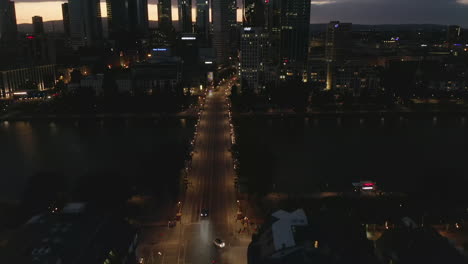 AERIAL:-Wide-Establishing-Shot-of-Frankfurt-am-Main,-Germany-Skyline-at-Night-over-Main-River-and-Bridge-with-Little-Car-traffic