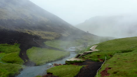 Aerial-view-of-Reykjadalur-valley-with-hot-springs-river-and-pool-with-lush-green-grass-meadow-and-hills-with-geothermal-steam.-Drone-view-of-bathing-in-thermal-hot-springs-in-Iceland