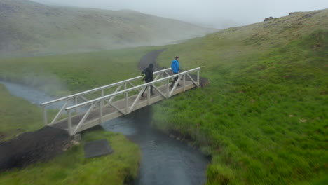 Drone-view-tourist-walking-footpath-in-Reykjadalur-Iceland-valley-in-foggy-day.-Birds-eye-of-hot-spring-steaming-pool-in-icelandic-grassy-countryside.-Natural-spa-in-Golden-Circle