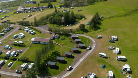 Directly-above-aerial-view-of-campsite-in-green-meadow-highlands-in-Iceland.-Top-down-view-of-characteristic-wooden-accommodation-bungalow-and-tourist-camping-in-motorhome