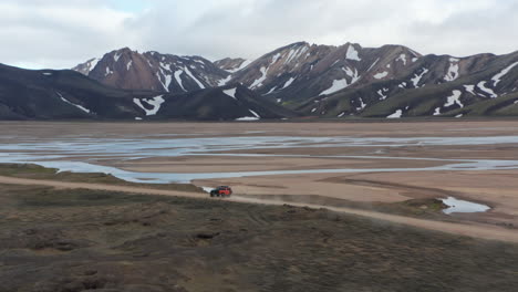 Drone-view-amazing-landscape-of-Icelandic-countryside-with-high-snow-mountain-peak.-Birds-eye-view-of-4x4-offroad-expedition-vehicle-speeding-driving-dirt-road-in-Iceland-highlands