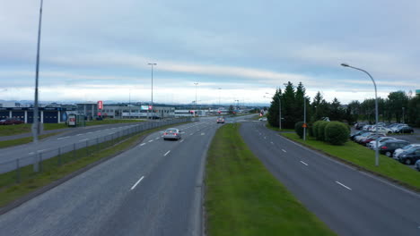 Forwards-fly-above-wide-multilane-trunk-road-with-intersection.--Cars-stopping-on-traffic-lights.-Green-lawn-space-along-road.-Reykjavik,-Iceland
