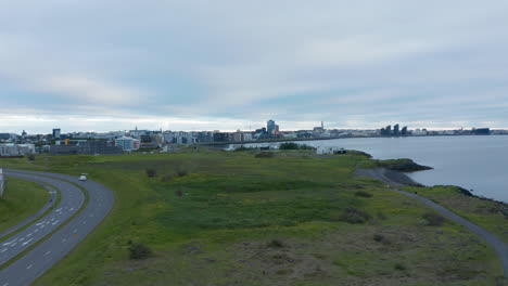 Elevated-shot-of-road,-green-meadow-and-sea-coast.-Building-in-city-on-bank-in-distance.-Cloudy-day.-Reykjavik,-Iceland