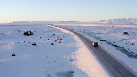 AERIAL:-Snow-White-Landscape-with-Road-following-Jeep-in-Iceland-at-Sunset-Winter,-Sun,-Arctic