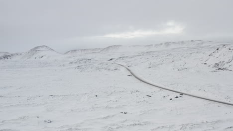 AERIAL:-Over-Snow-White-Landscape-with-Road,-Mountain-in-Iceland-Winter,-Snowing,-Cold,-Arctic