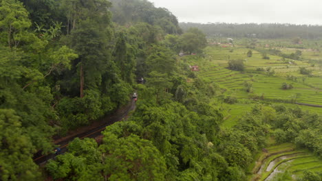 Aerial-view-of-vehicles-driving-through-rural-countryside-in-Bali-in-the-rain.-Cars-and-motorcycles-driving-on-the-road-near-tropical-rainforest-and-lush-green-rice-farm-fields