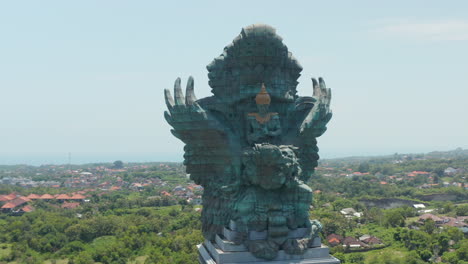 Front-rotating-aerial-view-of-majestic-copper-and-brass-Garuda-Wisnu-Kencana-statue-in-Bali,-Indonesia.-Large-blue-green-statue-rising-above-the-residential-homes