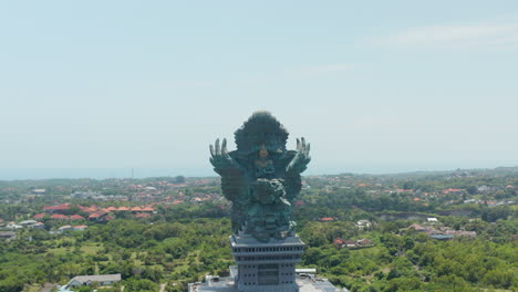 Giant-copper-statue-of-Garuda-Wisnu-Kencana-in-cultural-park-in-Bali,-Indonesia.-Aerial-dolly-retreating-view-of-large-religious-sculpture-rising-above-the-city