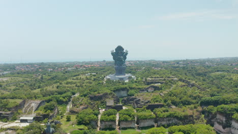 Large-statue-of-Garuda-Wisnu-Kencana-in-cultural-park-in-Bali,-Indonesia.-Forward-dolly-aerial-view-of-large-religious-monument-surrounded-by-thick-green-trees-in-a-park