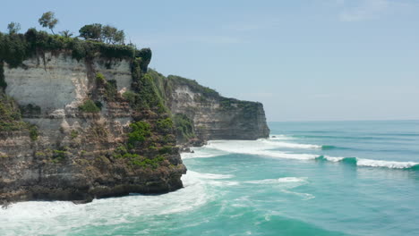 Stunning-tropical-coastline-and-blue-ocean-in-Bali,-Indonesia.-Aerial-view-of-large-waves-crashing-in-rocky-cliffs-in-Bali