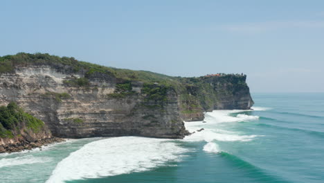 Small-homes-on-top-of-steep-ocean-cliffs-in-Bali,-Indonesia.-Aerial-view-of-large-steep-cliffs-and-beautiful-blue-ocean-in-Bali