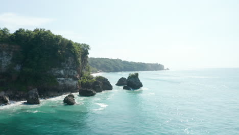 Low-aerial-shot-flying-by-steep-ocean-cliffs-above-tropical-turquoise-blue-sea-in-Bali,-Indonesia