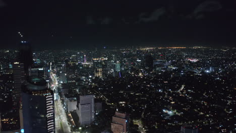 Aerial-wide-slow-dolly-shot-of-stunning-modern-city-center-at-night-with-skyscrapers-and-busy-multi-lane-highway-with-lights-in-Jakarta,-Indonesia
