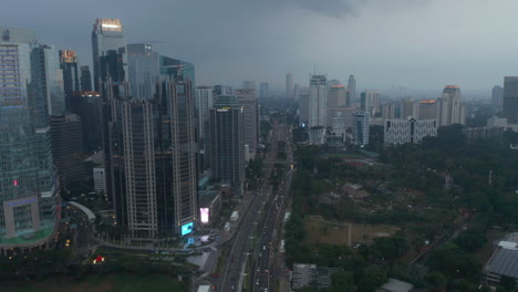 Luftwagenansicht,-Die-Abends-Dem-Verkehr-Auf-Einer-Stark-Befahrenen-Autobahn-Folgt,-Umgeben-Von-Hohen-Wolkenkratzern-Und-Einem-Sportplatz-In-Jakarta