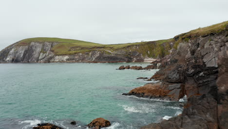 Fly-over-coastal-rocks.-Waves-washing-jagged-sea-coast.-Revealing-people-on-small-sand-beach-under-cliff.-Amazing-nature-scenery.-Ireland