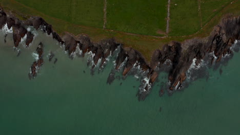 Aerial-birds-eye-overhead-top-down-view-of-waves-crashing-on-jagged-rocky-sea-coast.-dark-rocks-rising-above-sand-seabed.-Ireland