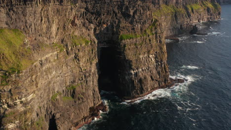 Flock-of-birds-flying-around-rock.-High-vertical-walls-of-cliffs-above-ripples-sea-lit-by-golden-hour-sunshine.-Cliffs-of-Moher,-Ireland