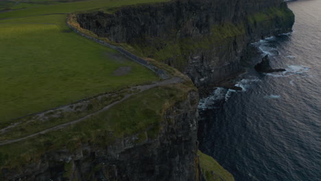 Aerial-ascending-footage-of-high-cliffs-on-sea-coast-at-dusk.-Tilt-down-on-panoramic-tourist-route-leading-on-edge-of-rock.-Cliffs-of-Moher,-Ireland