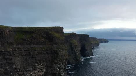Fly-along-rugged-rocky-sea-coast.-High-vertical-walls-above-rippled-water-surface-at-dusk.-Cliffs-of-Moher,-Ireland