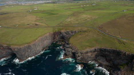 Aerial-panoramic-shot-of-green-grasslands-along-sea-coast.-High-and-jagged-rocky-cliffs-above-crashing-waves.-Kilkee-Cliff-Walk,-Ireland