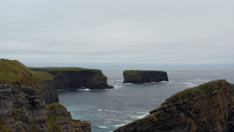 Forwards-fly-along-sea-coast.-High-cliffs,-rocky-islands-and-islets-in-waves.-Aerial-footage-on-natural-scenery.-Kilkee-Cliff-Walk,-Ireland