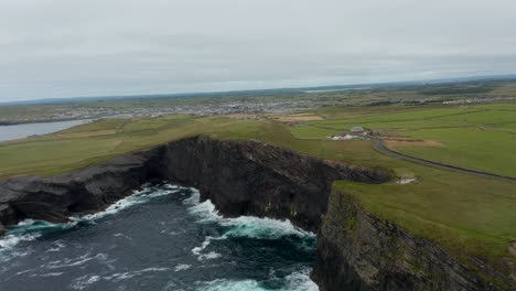 Aerial-ascending-shot-of-rough-waves-crashing-on-high-rocky-cliff.-Beautiful-natural-landmark.-Panoramic-view-of-flat-landscape.-Kilkee-Cliff-Walk,-Ireland