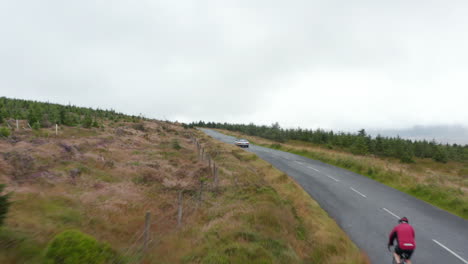 Cyclists-and-cars-on-mountain-road-leading-up-hill.-Forest-with-young-trees-along-road.-Overcast-sky.-Ireland