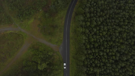 Aerial-birds-eye-overhead-top-down-panning-view-of-car-driving-on-road-through-forest.-View-through-sparse-clouds.-Ireland