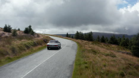 Silver-vintage-convertible-sports-car-with-textile-roof-on-road-winding-uphill-between-forest-and-meadows.-Ireland