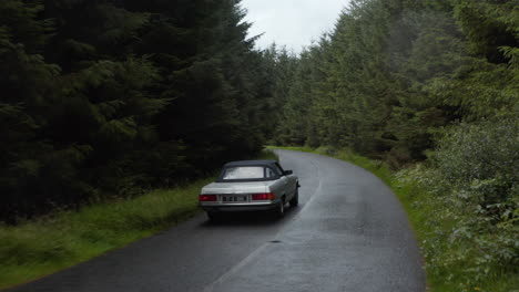 Forwards-tracking-of-vintage-sports-car-driving-on-narrow-wet-road-in-dense-forest.-Rainy-autumn-day.-Ireland