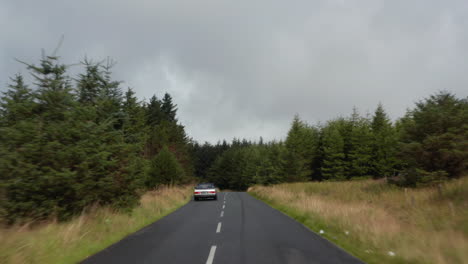 Forwards-tracking-of-vintage-car-driving-on-wet-road-surrounded-by-meadows-and-forest.-Braking-before-curve.-Ireland