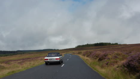 Low-flight-above-road-passing-through-large-moorlands.-Following-silver-vintage-sports-car-on-cloudy-day.-Ireland