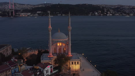 Ortakoy-Moschee-Beleuchtet-In-Schönem-Gelbem-Licht-Auf-Der-Wasserseite-In-Der-Abenddämmerung-Mit-Bosporus-Und-Brücke,-Luftaufnahme