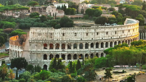 Aerial-descending-footage-of-worlds-largest-amphitheatre-in-golden-hour.-Ancient-Colosseum-and-surrounding-green-vegetation-in-parks.-Rome,-Italy