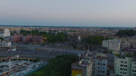 Slowly-moving-passenger-train-on-railway-tracks.-Forwards-fly-above-buildings-along-railroad-in-city-at-dusk.-Rome,-Italy