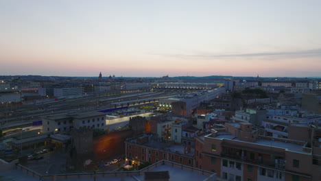 Aerial-view-of-illuminated-central-train-station-under-colour-twilight-sky.-Sliding-reveal-of-residential-buildings-in-city.-Rome,-Italy