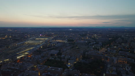 Aerial-panoramic-view-of-evening-city-and-colour-twilight-sky-in-background.-Richly-illuminated-central-train-station.-Rome,-Italy