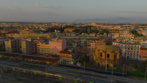 Forwards-fly-above-morning-city.-Commuter-train-driving-on-railway-tracks,-colour-buildings-in-urban-borough-Tempio-di-Minerva-Medica-historic-landmark.-Rome,-Italy