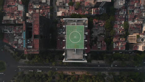 Aerial-birds-eye-overhead-top-down-view-of-tall-office-building-with-green-heliport-on-top-and-surrounding-area.-Camera-zooming-out.-Mexico-city,-Mexico.